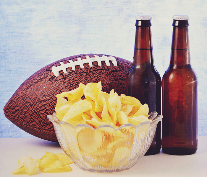 Two bottles of beer with a football and bowl of chips